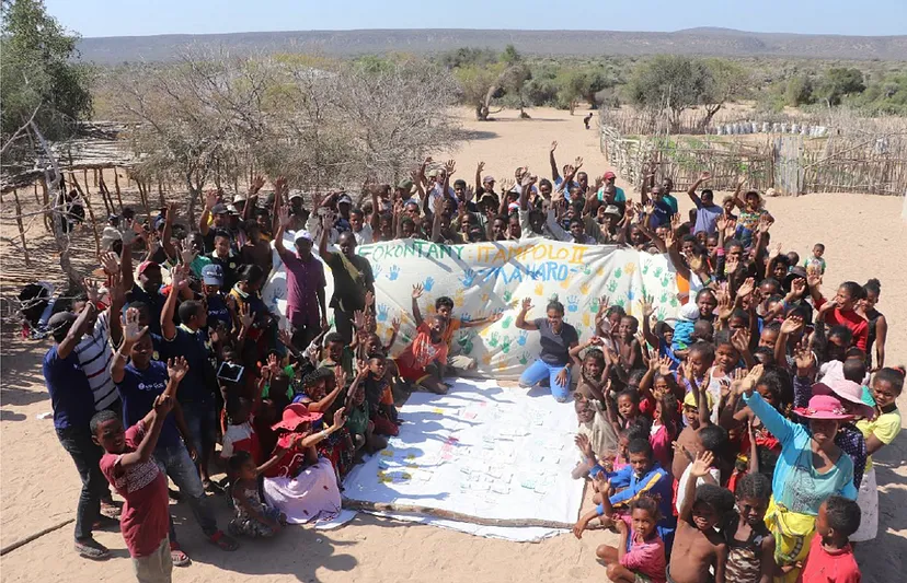 A group of children and adults raise their hands as they congregate around a sheet with handprints and painted text on it.