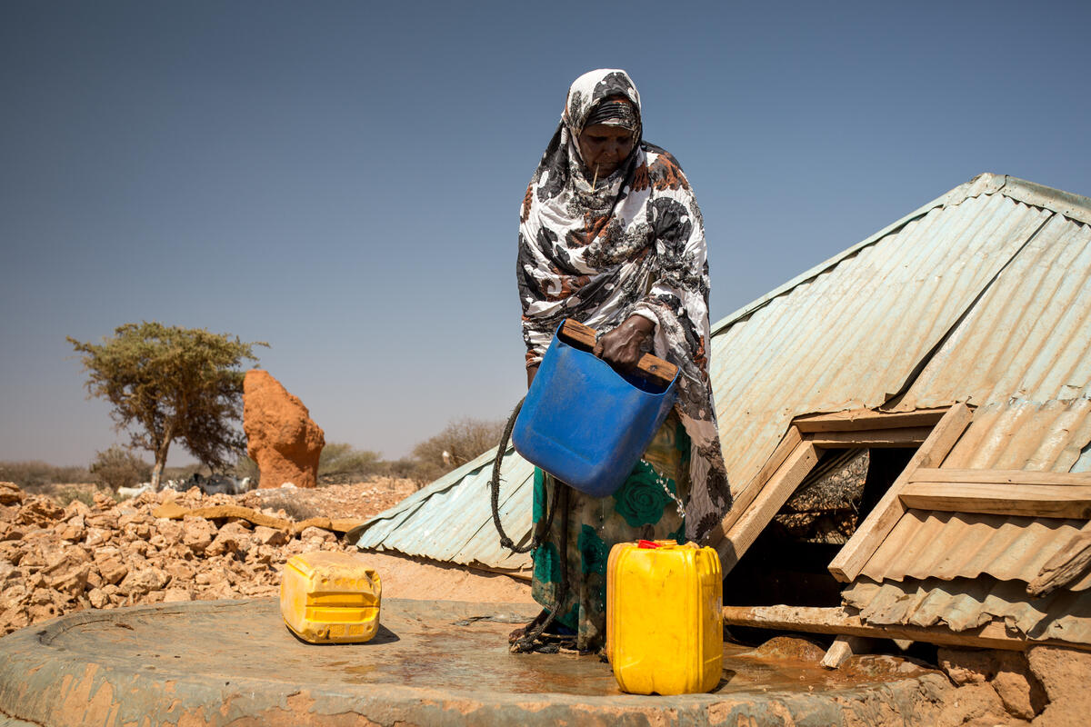 A woman pours water from a blue bucket into a yellow bidon.