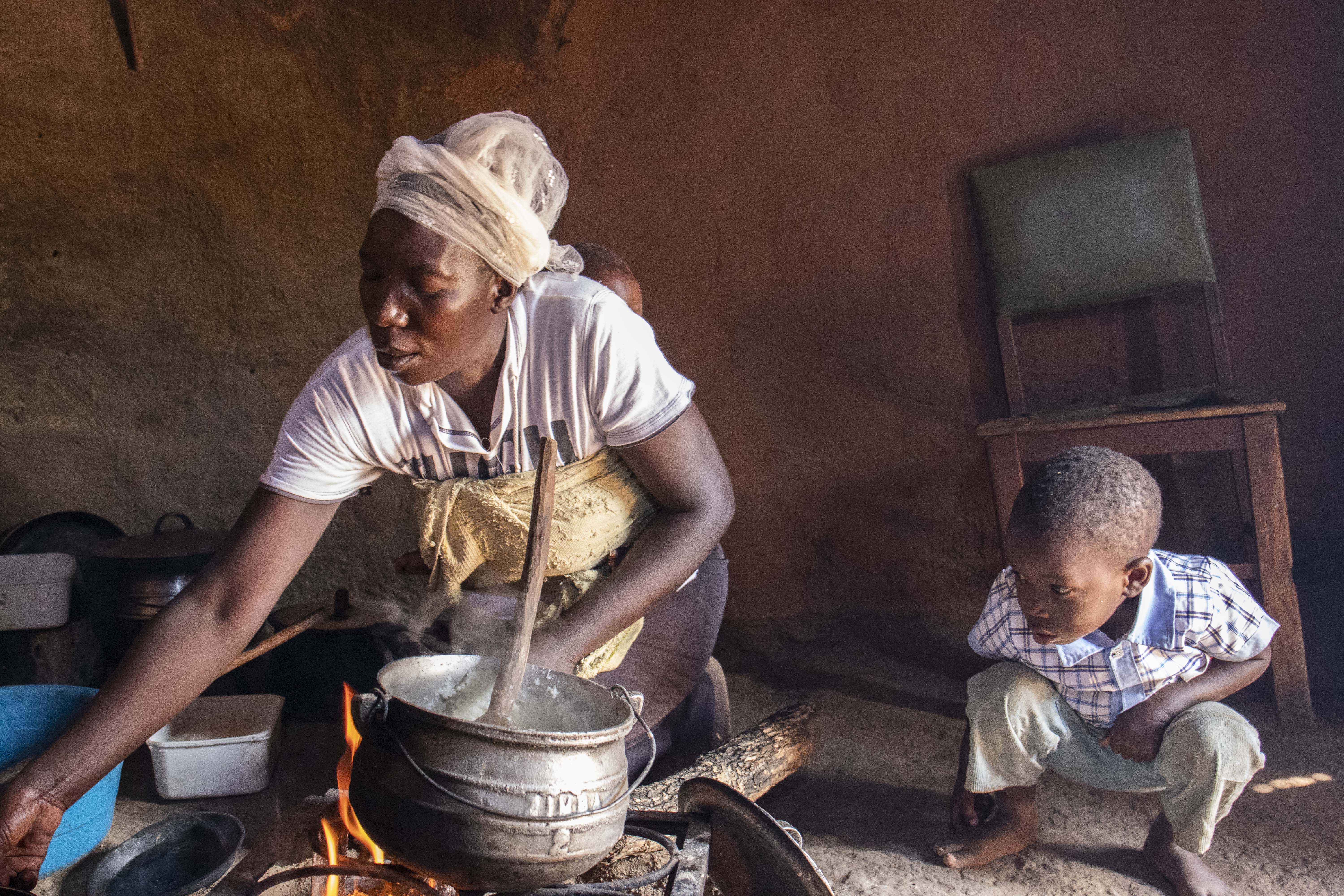 A woman carrying a baby on her back and young son watching, prepares food in a pot over an open fire, using food bought through a cash transfer, in Zimbabwe.