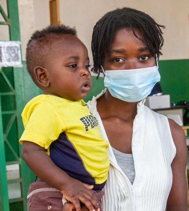 A woman wearing a facemask is holding a young child wearing a yellow shirt.