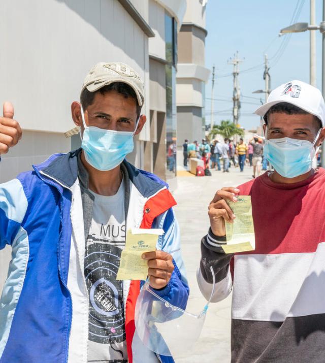 Two men wearing facemasks and baseball hats hold up cash assistance vouchers, and each raise a fist with a thumbs up
