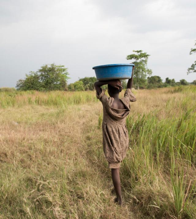 A young woman carries a bucket on her head as she walks through a field.