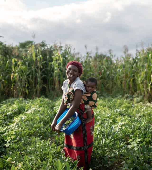 A woman holding a bucket and her child smiles while standing in a field of crops.