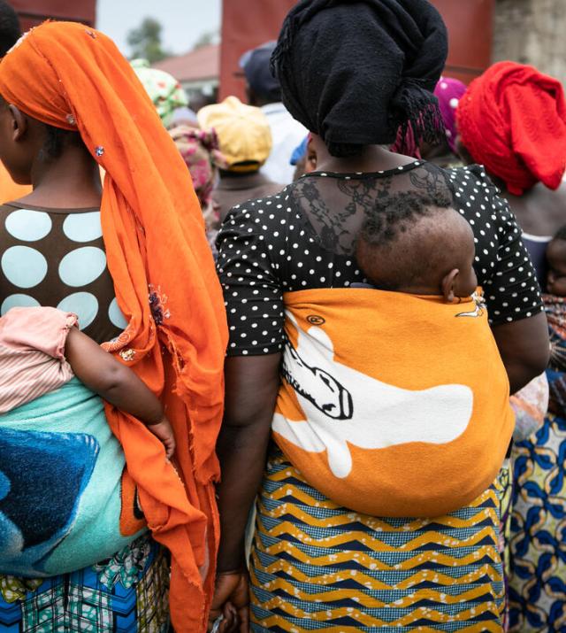 Three mothers with children strapped to their backs stand in line for supplies.