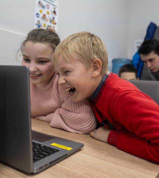 Two children work together at a laptop, laughing.