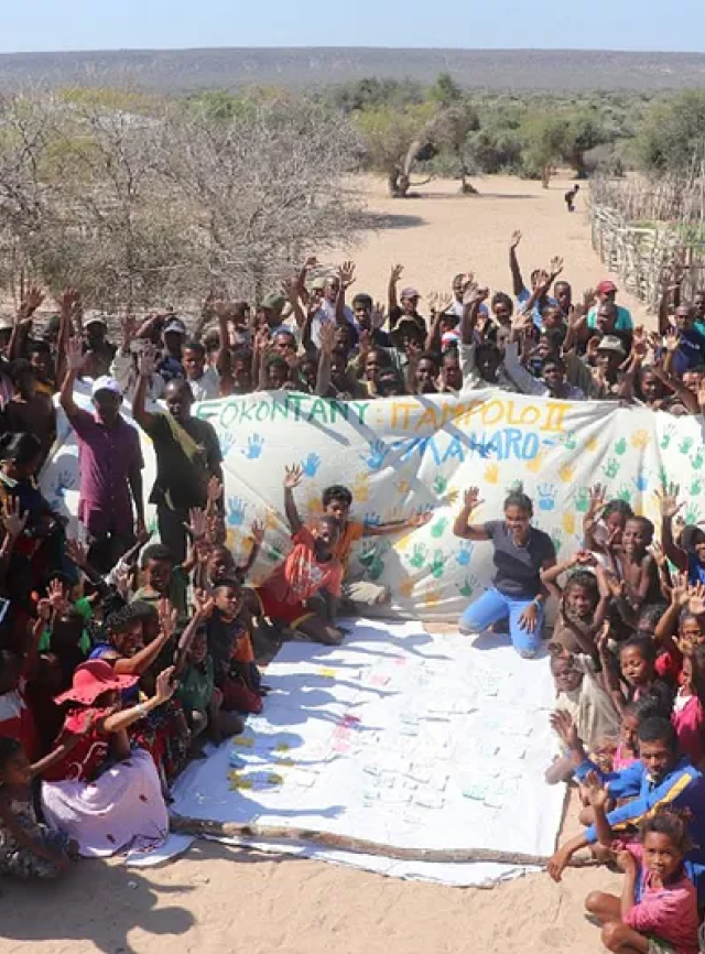 A group of children and adults raise their hands as they congregate around a sheet with handprints and painted text on it.