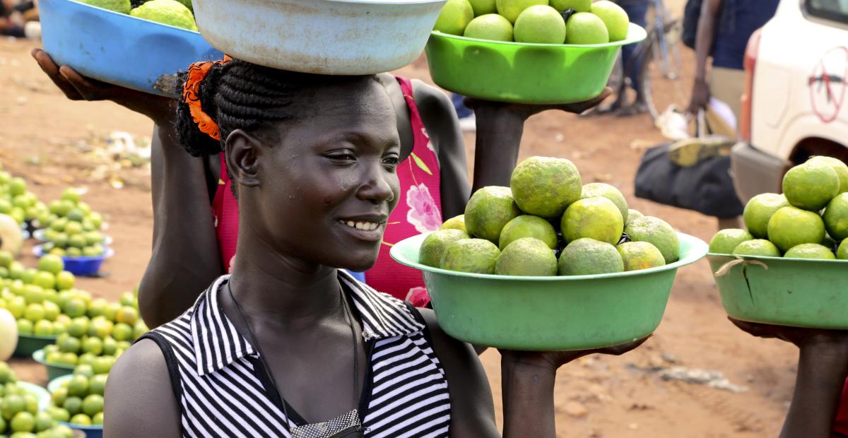 A woman selling green produce holds bowls with round green produce in her hand and on her head.