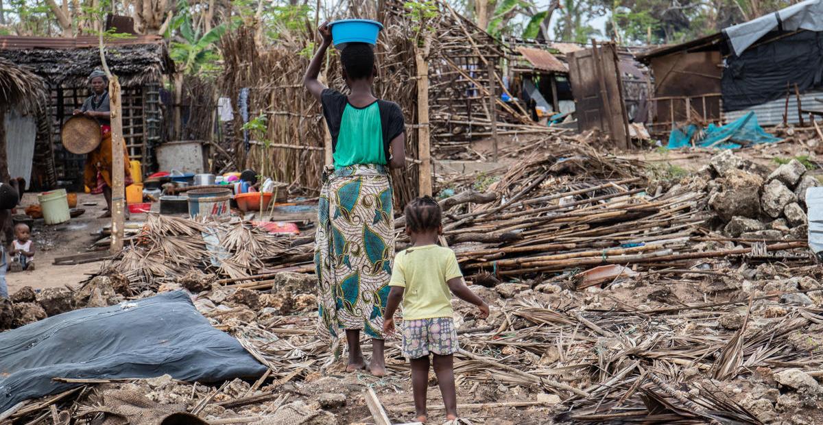 A woman carrying a blue bucket on her head walks with a small child through a damaged home in Mozambique.