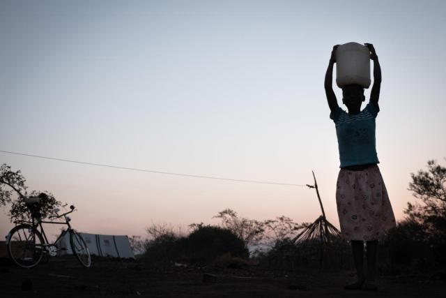 A young girl carries a jerry can of water on her head, the photo is in shadow with a sunset in the background.