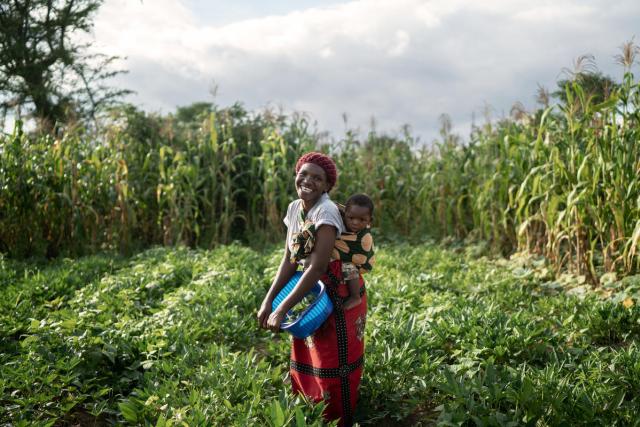 A woman holding a bucket and her child smiles while standing in a field of crops.