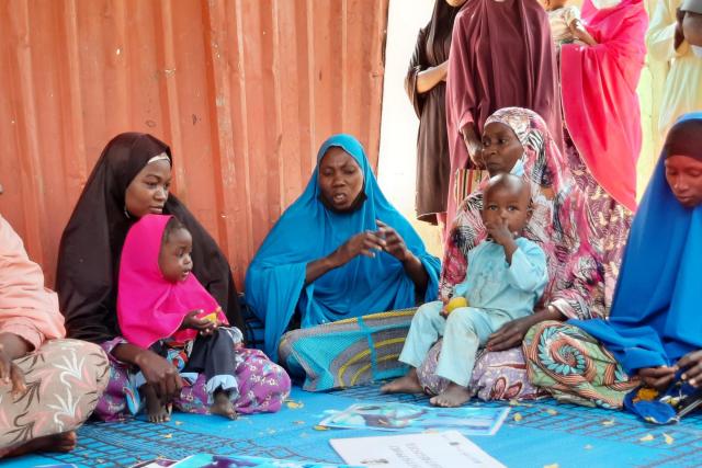 Four women holding two small children sit on a blue map in discussion.