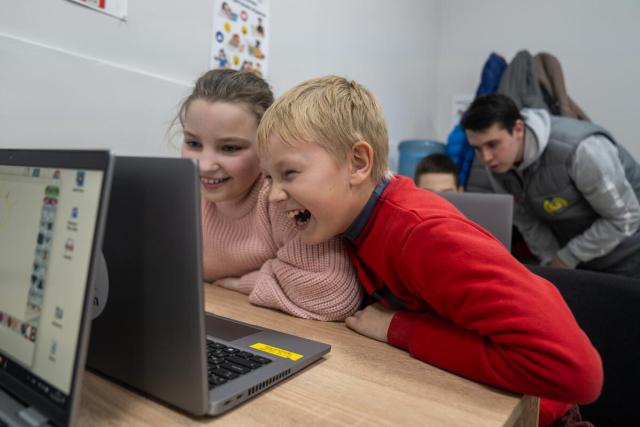 Two children work together at a laptop, laughing.