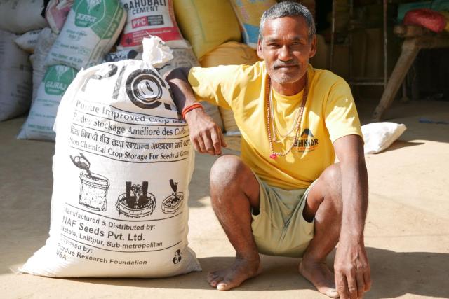 A person wearing a yellow shirt squats next to a large white bag of seeds.