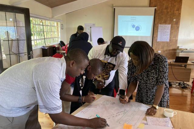 A group of people stand around a table writing on a large piece of paper during a workshop.